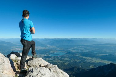 A man in hiking outfit with a panoramic view on the Alpine valley from the top of Mittagskogel in Austrian Alps. Clear and sunny day. There is a lake at the bottom. Outdoor activity. Achievement