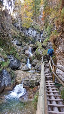 A man with blue backpack walking along a wooden, ladder pathway along a high gorge in Austrian Alps. There is a waterfall in front of her. Adventure park. Blue sky above. Autumn in the mountains.