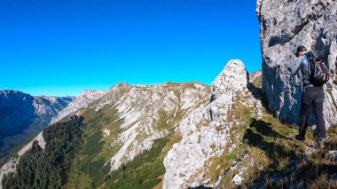A man with a hiking backpack climbing to the of a big boulder on the way to Hohe Weichsel in Austria, with a panoramic view on a vast valley.  Narrow pathway. He is enjoying the view. Discovering