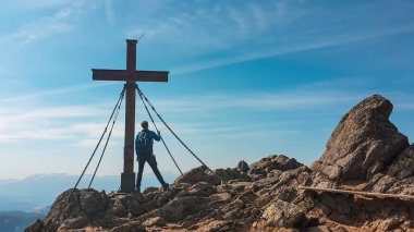 A man with hiking backpack standing on sharp rocks, under a wooden cross on top of Sauofen in Austrian Alps. A vast, golden pasture around. Fall vibes. Mountain chains in the back. Serenity