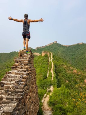 A man standing on top of a staircase of an unrenewed Gubeikou part of Great Wall of China, and spreading his arms. The wall is spreading on tops of mountains. Dense forest around it. World wonder.