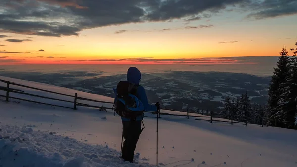 A man with hiking backpack and sticks standing at the snow covered peak of Schoeckl in Austrian Alps and admiring a sun rising above the horizon. The sky is bursting orange. Winter wonderland Day break