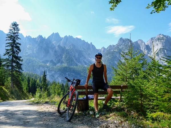 A man in biking outfit standing next to orange mountain bike next to a gravelled road in the mountains with the view on high Alps in the region of Gosau, Austria. Stony and barren mountain chains.