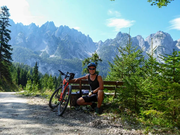 A man in biking outfit squatting next to orange mountain bike next to a gravelled road in the mountains with the view on high Alps in the region of Gosau, Austria. Stony and barren mountain chains.