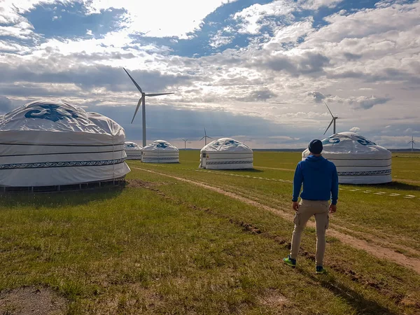A man standing in front of white, traditional yurts located on a pasture in Xilinhot in Inner Mongolia. Endless grassland. Blue sky with thick clouds. Wind turbines. Clean energy. Nomadic way of life