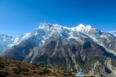 A panoramic view on steep Himalayan slopes along Annapurna Circuit Trek, Nepal. High, snow caped Annapurna peaks in the back catching the sunbeams. Serenity and calmness. Idyllic landscape.