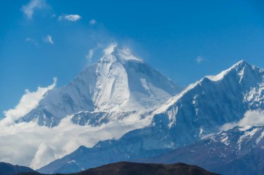 A distant view on snow capped Dhaulagiri I, seen from Mustang Valley, Annapurna Circuit Trek in Nepal. Wind blows the snow over the mountain peak. Barren and steep slopes. Harsh landscape.