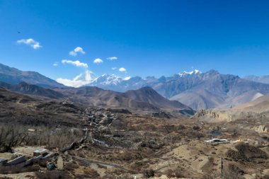 A small Himalayan village, Muktinath, along Annapurna Circuit Trek in Nepal. There is a pathway along a wall. There are only a few buildings. In the back there is high, snow capped Himalayan chain.