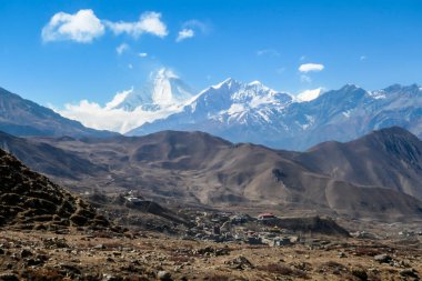 A small Himalayan village, Muktinath, along Annapurna Circuit Trek in Nepal. There is a pathway along a wall. There are only a few buildings. In the back there is high, snow capped Himalayan chain.