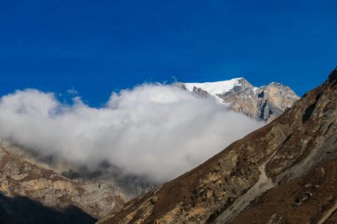 Thick clouds breaching through high mountain peaks in Muktinath, Annapurna Circuit Trek, Nepal. Clear blue sky. Snow capped mountains.