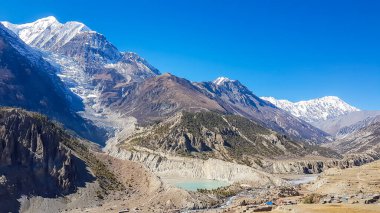 A panoramic view on Manang valley from Praken Gompa, Nepal. High Himalayan ranges around. There is a small lake in the valley. Snow capped peaks of Annapurna Chain. Harsh landscape.