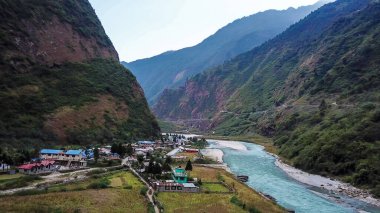 A turquoise river flowing at the bottom of Himalayan valley, surrounding small village Tal, Nepal. There is an Annapurna Circuit Trek going along the valley. Drone capture. High mountain chains