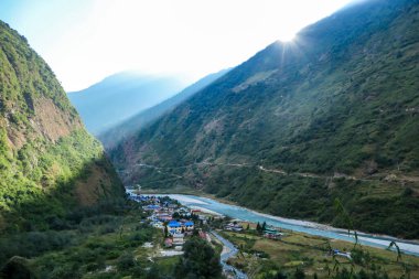 A turquoise river flowing at the bottom of Himalayan valley, surrounding small village Tal, Nepal. There is an Annapurna Circuit Trek going along the valley. Drone capture. High mountain chains