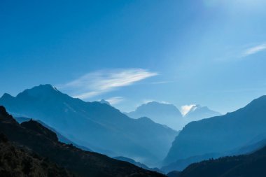 Himalayan chains shrouded in fog, seen from Thorung Phedi, Annapurna Circuit Trek, Nepal. There are multiple mountain chains. Sunbeams breaching through the peaks. Golden hour. Meditation and serenity