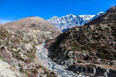 A view on rushing brook in Manang Valley, Annapurna Circus Trek, Himalayas, Nepal, with  Annapurna Chain and Gangapurna in the back. Dry and desolated landscape. High, snow capped mountain peaks.
