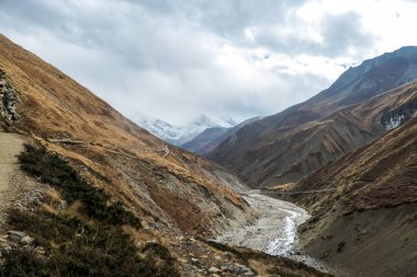 A view on rushing brook in Manang Valley, Annapurna Circus Trek, Himalayas, Nepal, with  Annapurna Chain and Gangapurna in the back. Dry and desolated landscape. High, snow capped mountain peaks.