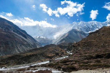 Panoramic view on a valley along Annapurna Circuit in Nepal, with a small torrent flowing in the middle. In the back there are high, snow capped Himalayan peaks. Slopes are overgrown with small bushes