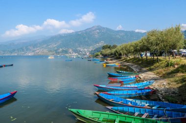 A view on Phewa Lake in Pokhara, Nepal with many colorful boats parked along it's shore. There are high Himalayan ranges in the back. Calm surface of the lake. Clear and sunny day. Serenity