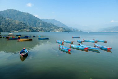 A view on Phewa Lake in Pokhara, Nepal with many colorful boats anchored around the shore. There are high Himalayan ranges in the back. Calm surface of the lake. Clear and sunny day. Serenity