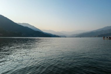 An idyllic view on Phewa Lake in Pokhara, Nepal. There are high Himalayan ranges around the lake. Calm surface of the lake. Clear and sunny day. There is a dence forest at the shore. Golden hour haze