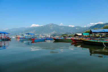 A view on Phewa Lake in Pokhara, Nepal with many colorful boats parked along it's shore. There are high Himalayan ranges in the back. Calm surface of the lake. Clear and sunny day. Serenity