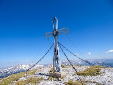 A cross on the top of Hochturm, Austrian Alps. The cross is supported by 4 metal ropes. Dense forest on both sides. Alpine landscape on a sunny, summery day. Exploring the nature