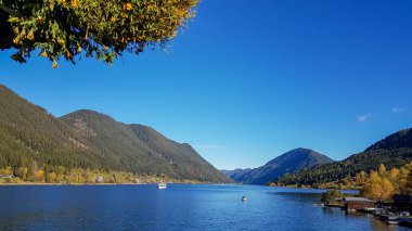 An idyllic view on the Weissensee lake surrounded by the Austrian Alps. The surface of the lake is calm and reflecting the mountains. Joyfulness and happiness. A tree crown in the upper corner