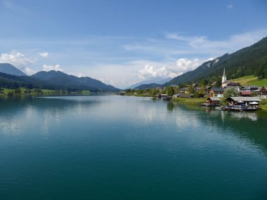 An idyllic, panoramic view on the Weissensee lake in Austria. The lake is surrounded by high Alps. There is a small village at the lake's shore, with tall church tower. Few clouds. Calmness and peace