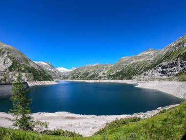 A panoramic view on an artificial, dam lake stretching over a vast territory around Alps in Austria. Lake is shining with navy blue color. In the back there are a few glaciers. Controlling the nature