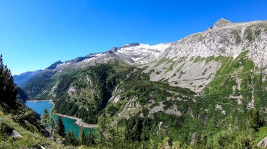 A panoramic view on an artificial, dam lake stretching over a vast territory around Alps in Austria. Lake is shining with navy blue color. In the back there are a few glaciers. Controlling the nature