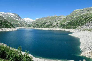 Dam in Austrian Alps. The artificial lake stretches over a vast territory, shining with navy blue color. The dam is surrounded by high mountains. In the back there is a glacier. Controlling the nature