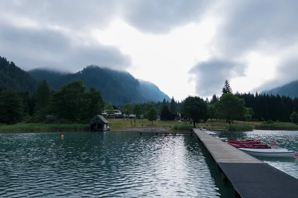 A camping place located at the shore of Weissensee lake surrounded by the Austrian Alps. The camping has a small pier, with some boats parked along. A bit of overcast. Camping in the nature