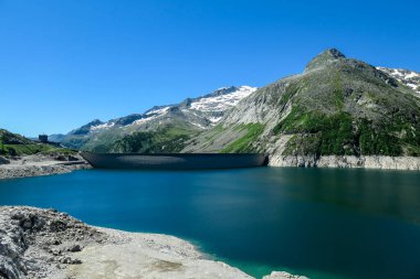 Dam in Austrian Alps. The artificial lake stretches over a vast territory, shining with navy blue color. The dam is surrounded by high mountains. In the back there is a glacier. Controlling the nature