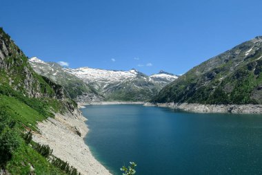 Dam in Austrian Alps. The artificial lake stretches over a vast territory, shining with navy blue color. The dam is surrounded by high mountains. In the back there is a glacier. Controlling the nature