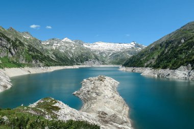 Dam in Austrian Alps. The artificial lake stretches over a vast territory, shining with navy blue color. The dam is surrounded by high mountains. In the back there is a glacier. Controlling the nature