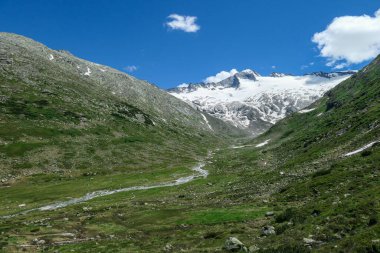 Panoramic view on huge glacier in Maltal region in Austrian Alps. The valley below it flourishing with green and freshness., small torrent in the middle. Summer in the mountains. Serenity and solitude