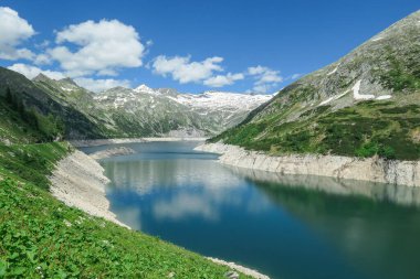 Dam in Austrian Alps. The artificial lake stretches over a vast territory, shining with navy blue color. The dam is surrounded by high mountains. In the back there is a glacier. Controlling the nature