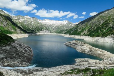 A torrent rushing towards a lake. The artificial lake stretches over a vast territory, shining with navy blue color. The dam is surrounded by mountains.A glacier in the back. Controlling the nature
