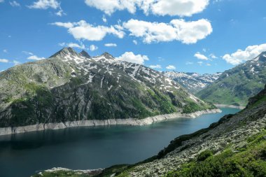 Dam in Austrian Alps. The artificial lake stretches over a vast territory, shining with navy blue color. The dam is surrounded by high mountains. In the back there is a glacier. Controlling the nature