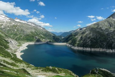 Dam in Austrian Alps. The artificial lake stretches over a vast territory, shining with navy blue color. The dam is surrounded by high mountains. In the back there is a glacier. Controlling the nature