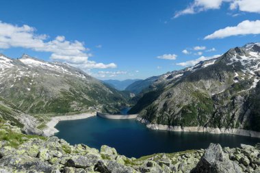 Dam in Austrian Alps. The artificial lake stretches over a vast territory, shining with navy blue color. The dam is surrounded by high mountains. In the back there is a glacier. Controlling the nature