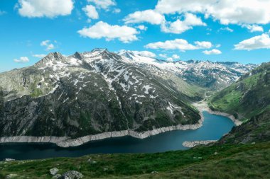 Dam in Austrian Alps. The artificial lake stretches over a vast territory, shining with navy blue color. The dam is surrounded by high mountains. In the back there is a glacier. Controlling the nature