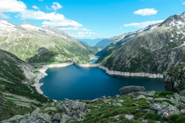 Dam in Austrian Alps. The artificial lake stretches over a vast territory, shining with navy blue color. The dam is surrounded by high mountains. In the back there is a glacier. Controlling the nature