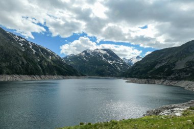 Dam in Austrian Alps. The artificial lake stretches over a vast territory, shining with navy blue color. The dam is surrounded by high mountains. In the back there is a glacier. Controlling the nature