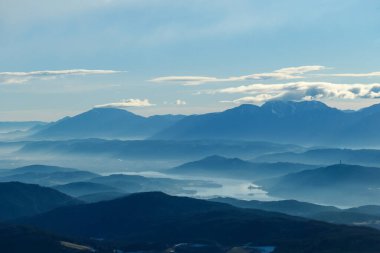 A panoramic view on Austrian Alps, captured on an early morning. The high peaks are hiding behind light clouds. The valley is shrouded in fog. There is a long lake at the bottom of the valley. Remedy