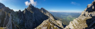 A panoramic view from an Alpine top on a vast valley. There are sharp mountains and high peaks around. The Alpine slopes are almost barren. Lush green valley. Bright day. Serenity and freedom.