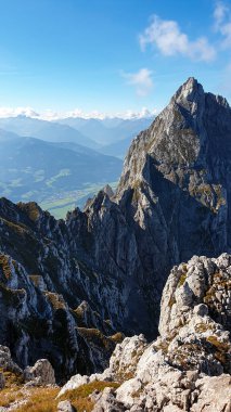 A panoramic view from an Alpine top on a vast valley. There are sharp mountains and high peaks around. The Alpine slopes are almost barren. Lush green valley. Bright day. Serenity and freedom.