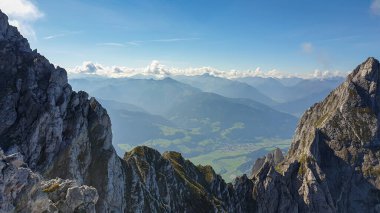 A panoramic view from an Alpine top on a vast valley. There are sharp mountains and high peaks around. The Alpine slopes are almost barren. Lush green valley. Bright day. Serenity and freedom.