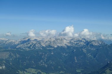 A panoramic view on a distant chain of Alps from another high peak. The massive chain is partially shrouded with clouds. Below there is lush green valley and a few cities in it. Austrian landscape