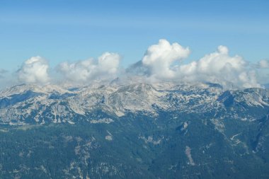A panoramic view on a distant chain of Alps from another high peak. The massive chain is partially shrouded with clouds. Below there is lush green valley and a few cities in it. Austrian landscape